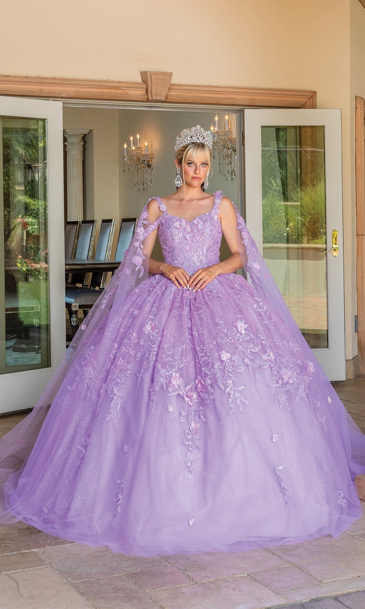 A person stands outside double doors wearing Quinceanera Dress 1664 By Dancing Queen, a light purple ball gown with floral details, lace embroidered cape sleeves, and a silver crown.